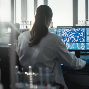 Medical Science Laboratory with Diverse Team of Professional Biotechnology Scientists Developing Drugs, Female Biochemist Working on Computer Showing Gene Therapy Interface. Back view Shot