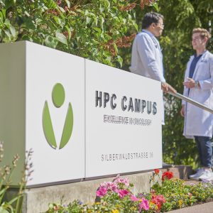 Zwei medizinische Fachkräfte in weißen Kitteln stehen im Gespräch neben einem modernen Eingangsschild mit der Aufschrift „HPC Campus – Excellence in Oncology“. Das Schild ist von Blumen und Grünpflanzen umgeben, im Hintergrund sind Bäume und eine Außentreppe zu sehen. Two medical professionals wearing white lab coats are talking next to a modern entrance sign reading “HPC Campus – Excellence in Oncology.” The sign is surrounded by flowers and greenery, with trees and an outdoor stairway visible in the background.