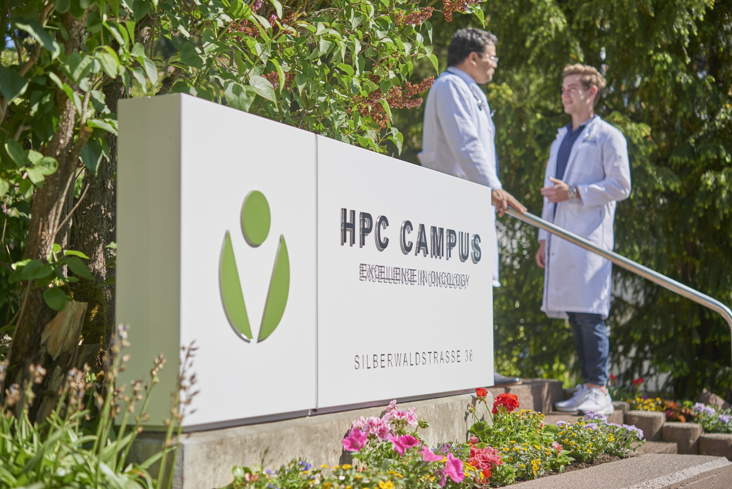 Zwei medizinische Fachkräfte in weißen Kitteln stehen im Gespräch neben einem modernen Eingangsschild mit der Aufschrift „HPC Campus – Excellence in Oncology“. Das Schild ist von Blumen und Grünpflanzen umgeben, im Hintergrund sind Bäume und eine Außentreppe zu sehen. Two medical professionals wearing white lab coats are talking next to a modern entrance sign reading “HPC Campus – Excellence in Oncology.” The sign is surrounded by flowers and greenery, with trees and an outdoor stairway visible in the background.